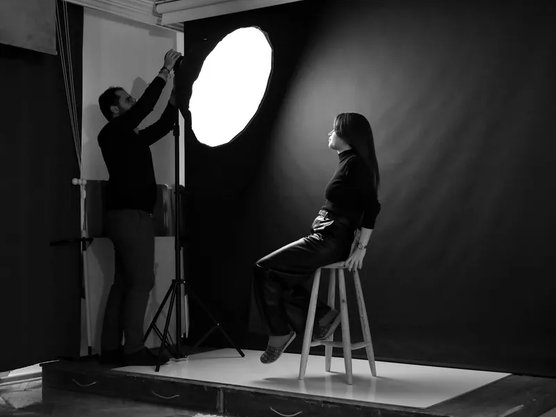 Black-and-white studio portrait session: a model on a stool while the photographer adjusts a light